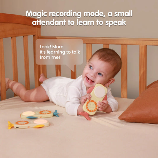 A baby lying in a wooden crib, smiling and holding a whale-shaped baby toy phone with pink fins, while two more toy phones are on the bed. Text above reads “Magic recording mode, a small attendant to learn to speak,” and a speech bubble says, “Look! Mom, It’s learning to talk from me!”