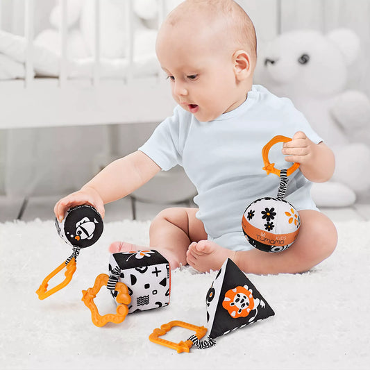 A baby sitting on a soft carpet, playing with black and white high-contrast soft toys in various shapes (ball, cube, pyramid, and cylinder). Each toy features an orange clip for easy attachment, designed to support infant visual development, sensory exploration, and grasping skills.