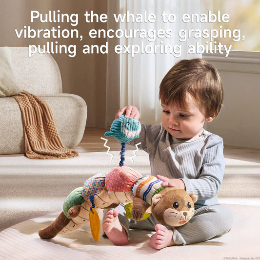 A toddler in light gray ribbed clothing sits on a cream-colored carpet, engaging with a multifunctional otter sensory toy. The child is pulling on the blue whale teether that extends on a coiled cord from the toy's body. The plush toy features a cute brown otter head with whiskers and eyes on one end, and a tail on the other, with five colorful segments in between (green dotted, multicolored squares, pink with circles, and striped patterns). Yellow leaf-shaped teethers hang from the toy. Text overlay at the