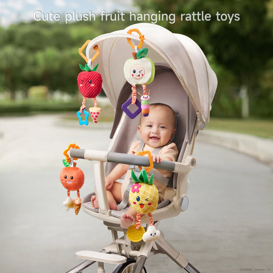 A smiling baby sitting in a stroller is surrounded by colorful plush fruit-themed hanging rattle toys—strawberry, apple, orange, and pineapple—each attached by orange hooks. The toys have cheerful embroidered faces and soft dangling elements like teething shapes and plush accessories. The background features a sunny, outdoor park pathway.