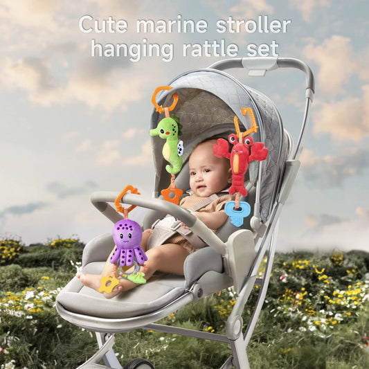 Baby sitting in a stroller outdoors, smiling and playing with a cute marine-themed hanging rattle toy set that includes a red lobster, purple octopus, and green seahorse plush toys, each attached to the stroller canopy with orange clips.