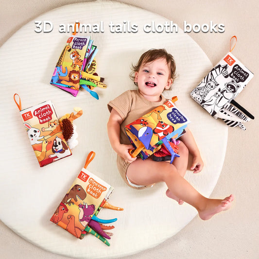 Smiling toddler sitting on a soft white mat, surrounded by colorful 3D animal tails cloth books. The child is holding the Marine Cloth Book and is surrounded by four other titles: Dinosaur Cloth Book, Forest Cloth Book, Animal Cloth Book, and a black-and-white Animal Cloth Book. Each book features soft fabric pages and textured animal tails sticking out, designed for sensory play and early learning.