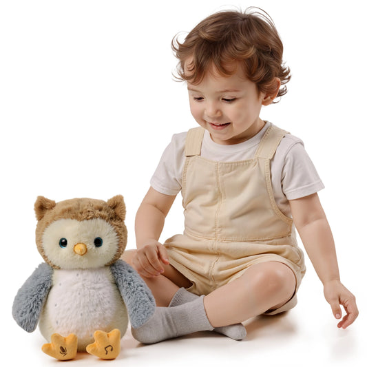 Smiling toddler sitting on the floor next to an interactive talking animated owl plush toy, which features soft brown and gray fur, a yellow beak, and orange feet with embroidered microphone and music note icons, indicating sound and music functions.