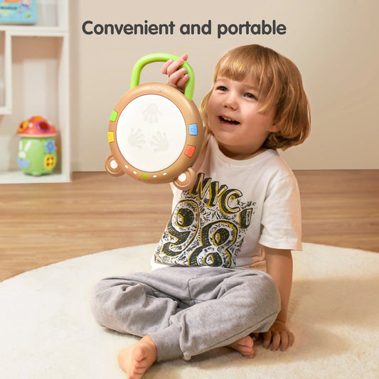 A smiling toddler sits on a soft rug in a playroom, holding a portable bear-shaped baby drum toy by its green handle. The drum features colorful buttons around the edges and handprint icons on the center surface. The text "Convenient and portable" is displayed above, highlighting the toy's easy-to-carry design.