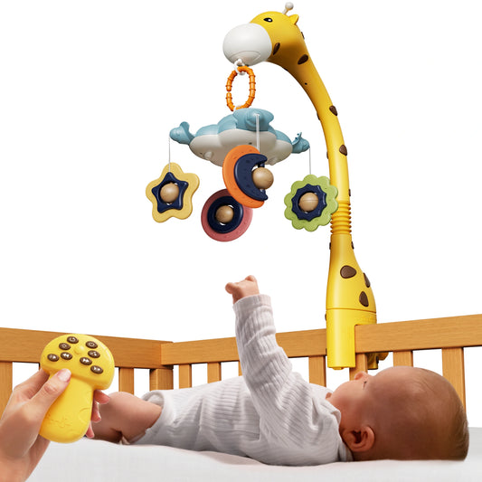 A baby lying in a wooden crib reaches up toward a giraffe-shaped crib mobile. The mobile hangs colorful star and moon toys with small balls inside. A hand is shown holding a yellow remote control with multiple buttons, used to operate the mobile’s music and motion features. The baby looks engaged and is stretching a hand upward toward the hanging toys.