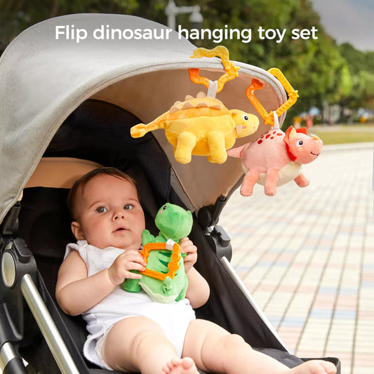 Baby sitting in a stroller reaching for three reversible plush dinosaurs—green, yellow, and pink—hanging from the canopy by bright orange clips