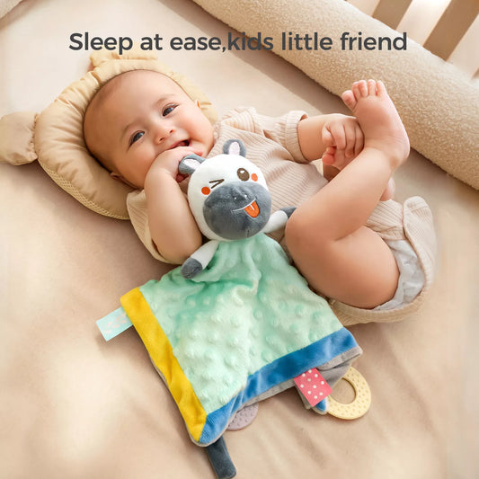 Smiling baby lying on a bed while holding a plush animal security blanket lovey with a green, yellow, and blue soft fabric. The toy has a cute winking animal face and attached teether corners.