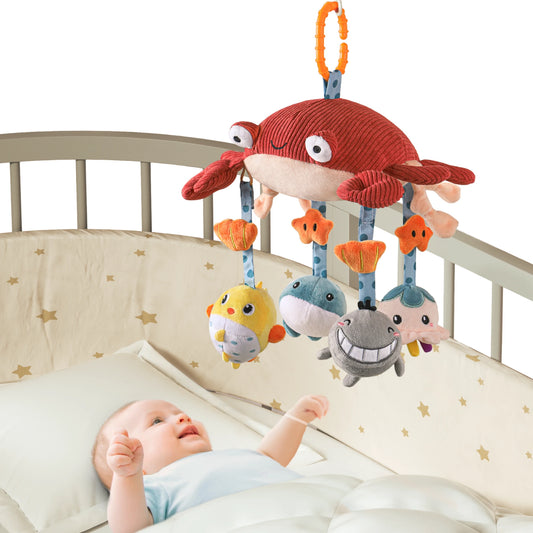 A cozy baby room features a crib with a white bedsheet patterned with gold stars. Suspended above the crib is a mobile from which hangs a red crab plush toy sporting a blue band around its arms on a blue, anchor-shaped hanger. Below the crib, three fish-shaped plush toys also hang—two in yellow with orange fins and one in blue with a white belly and smiling face. A baby lies on the bed, facing the crib and reaching out toward the crab toy.