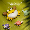 A yellow crab-shaped bubble bath toy is placed on a green surface along with three smaller bath toys: a purple penguin, a red crab, and a green whale. Small white pellets are scattered around, emphasizing the durable design. The text “High quality and safe ABS materials” appears at the top, highlighting the toy’s child-safe construction.