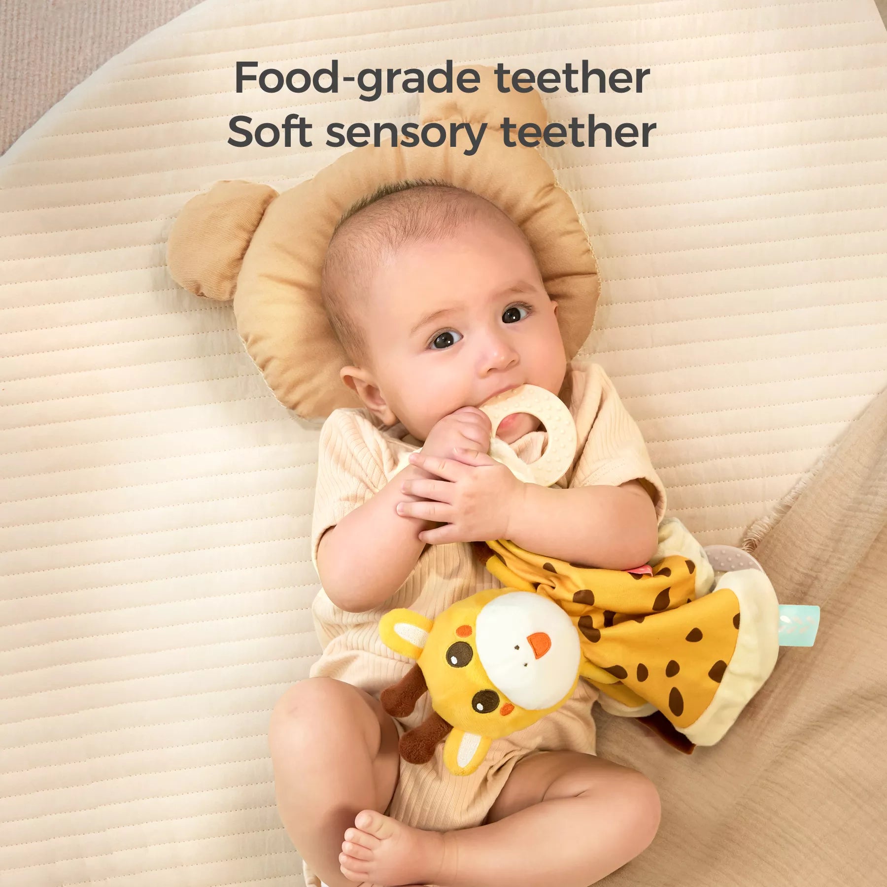 A baby lying on a bed with a bear-shaped pillow, holding and chewing on a food-grade teether attached to a giraffe baby security blanket. The text above says, "Food-grade teether, Soft sensory teether."