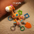 Colorful plush giraffe baby toy lying on a soft beige carpet next to a brown pillow. The toy features various textures, crinkle elements, and attached teething rings in different shapes and colors, including a green star, blue square, orange fish, and clear rattle ring.