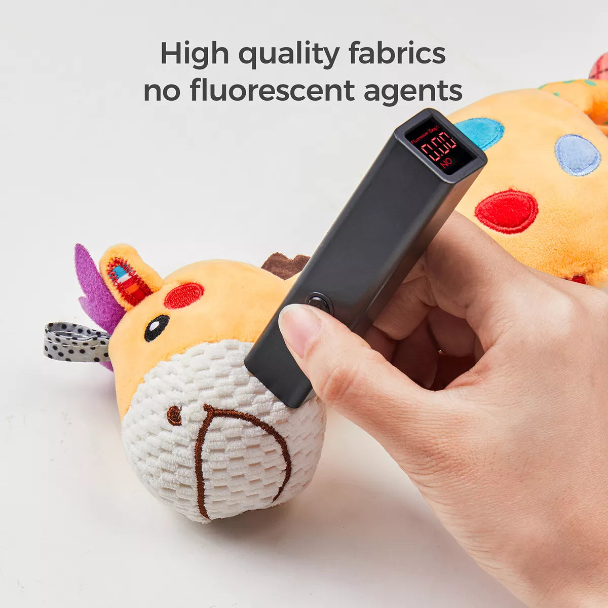 Close-up of a hand using a black UV detector to scan the plush giraffe toy’s face. The screen reads “NO,” indicating no fluorescent agents detected. Text at the top says “High quality fabrics no fluorescent agents,” highlighting the toy's safety and non-toxic materials.