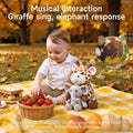 A baby sitting on a picnic blanket outdoors, playing beside a plush giraffe and elephant toy duo that performs musical interactions. Text on the image highlights the giraffe singing and the elephant responding, with a small inset showing the giraffe playing music.
