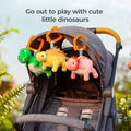 Three reversible plush dinosaur hanging toys—a green T‑rex, yellow stegosaurus, and pink triceratops—clipped to the canopy of a gray stroller outdoors