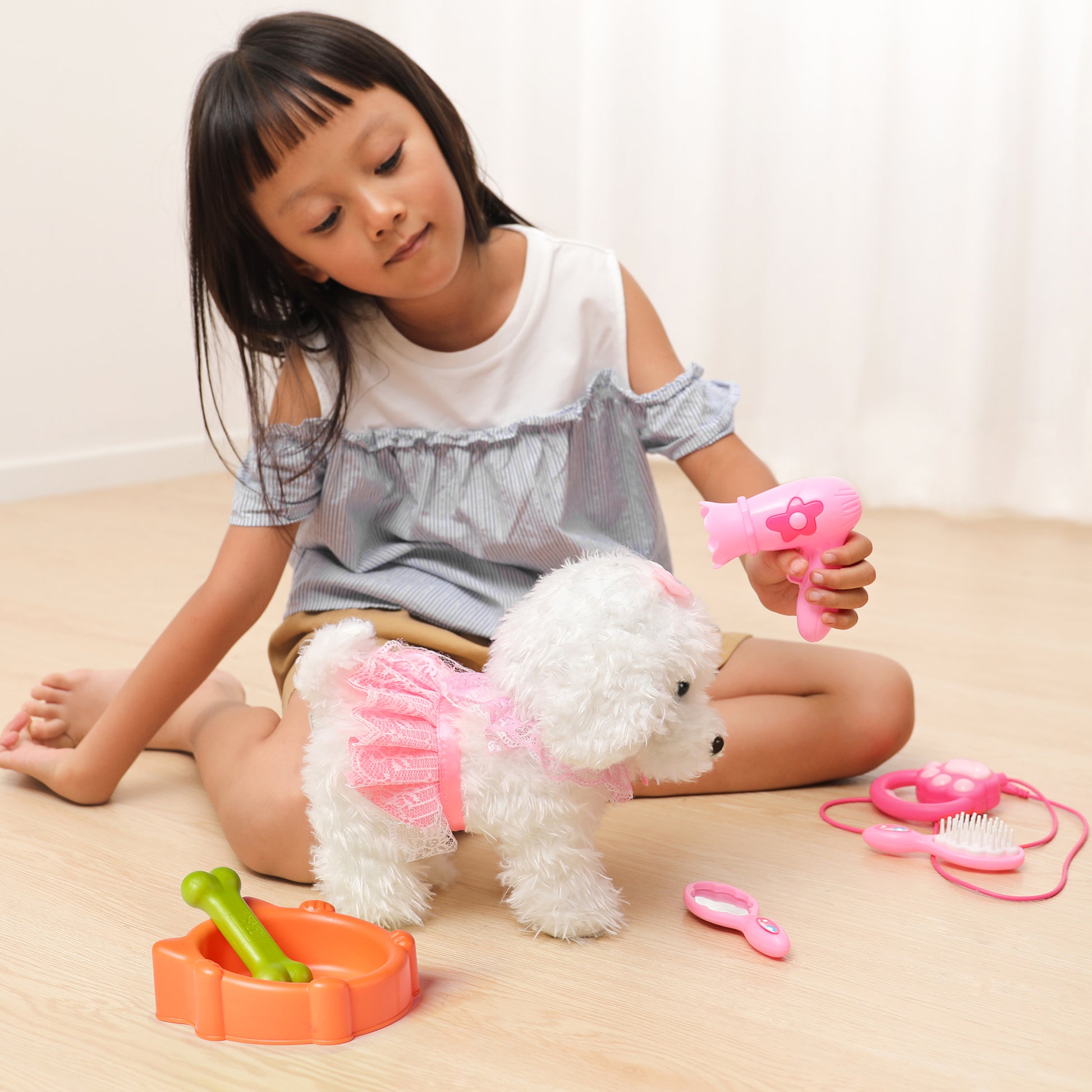 Young girl sitting on the floor playing with a white plush dog toy in a pink lace dress, holding a toy hairdryer as part of a pretend pet grooming set, with accessories like a brush, mirror, stethoscope, and dog bowl nearby.