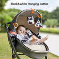 A baby lying in a stroller outdoors, playing with black and white high-contrast hanging rattle toys in various shapes. The toys are attached to the stroller canopy with orange clips, promoting visual stimulation, auditory development, and motor skills during travel or walks.
