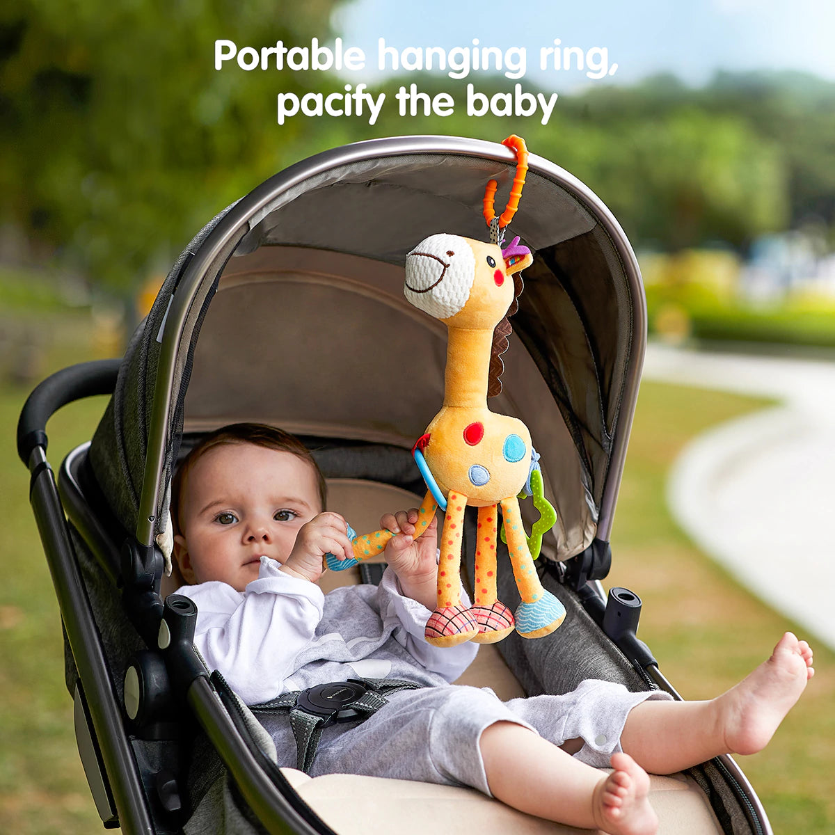 Baby sitting in a stroller outdoors, holding and playing with a plush giraffe toy hanging from the stroller canopy using an orange clip ring. The giraffe toy features colorful spots, teething rings, and textured legs. Text at the top reads “Portable hanging ring, pacify the baby,” emphasizing its calming and on-the-go use.