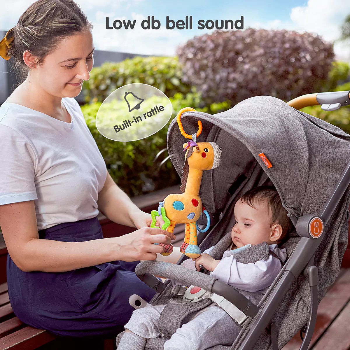 A woman plays with a plush giraffe toy to engage a baby seated in a gray stroller. The giraffe is hanging from the stroller canopy by an orange clip ring and features teething accessories and soft textures. On-screen text reads “Low db bell sound attract” and “Built-in rattle,” highlighting its gentle sound feature for sensory stimulation.